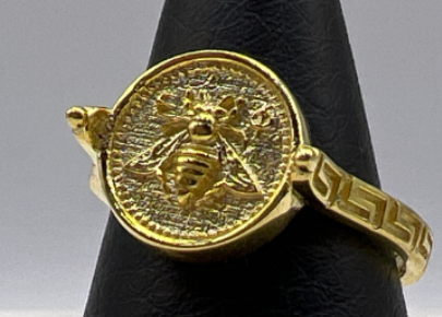 Gold ring with bee design on a black stand against a gray background